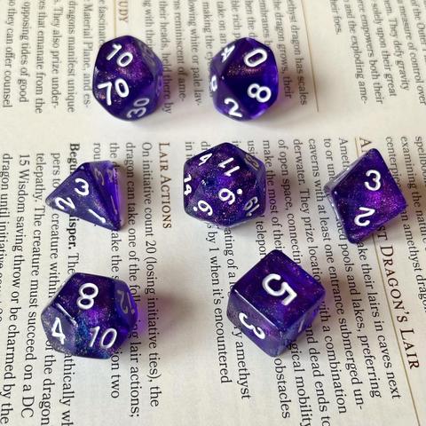 Picture of a set of transparent royal purple polyhedral dice with white numbers & green-blue-purple-gold glitter inside, sitting on a page from the D&D book Fizban’s Treasury of Dragons.