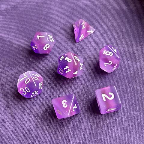 Picture of a set of translucent purple polyhedral dice with white numbers, 4 layers of different shades of purple, & subtle silver glitter throughout, sitting on a darker purple background with a denim texture.