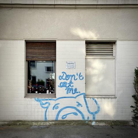 auf dem bild ist eine weiße hauswand mit zwei fenstern zu sehen. auf der wand befindet sich ein blaues graffiti eines traurigen schweins mit dem schriftzug „don’t eat me.“ darüber hängt ein schild mit dem text „privatparkplatz“. links neben dem graffiti ist ein fenster mit holzjalousie und mehreren flaschen auf der fensterbank zu sehen. das bild wirkt wie eine stille botschaft gegen fleischkonsum.
