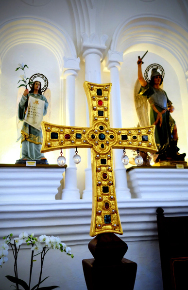 In einer Kirche steht im Altarbereich ein goldenes Kreuz, das mit Schmucksteinen besetzt ist. Im Hintergrund stehen in Nischen neben Säulen Heiligenstatuen, u.a. des Namenspatrons des Dorfes.