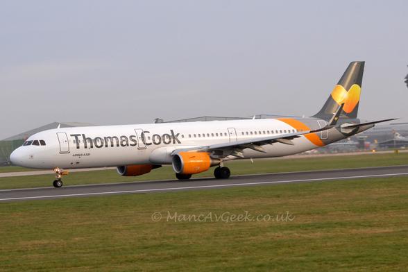 Side view of a white, twin engined jet airliner moving from left to right on a black runway.
The plane is mostly white, with grey, billboard-style "Thomas Cook" titles on the forward fuselage, withn a 2-tone grey tail with a diagonal orange stripe on the rear fuselage, and a yellow and orange love heart on the tail.
The engine pods are a bright orange.
Green grass fills the foreground, with more grass on the other side of the runway visible under the plane. The roofs of a couple of grey hangars are visible over the top of the plane, under grey sky.