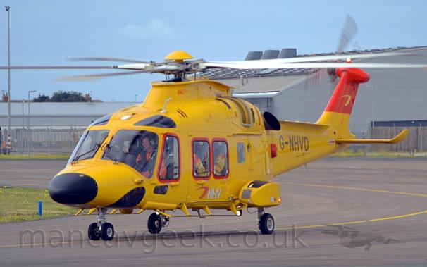 Side view of a yellow helicopter taxiing from right to left.
The  helicopter has a black nose-cone, and a white and red "NHV" logo on the side of the body, under the cabin windows.
The tip of the tail is bright red.
The tail-boom has the black registration "G-NHVD".
Grey taxiways fill the foreground and background, with grey buildings in the distance, under blue-grey skies.