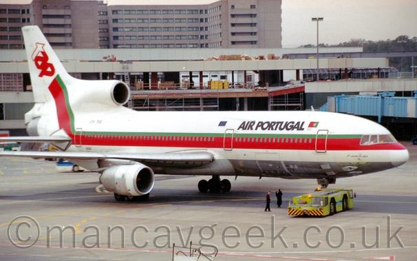 Side view of a white, 3 engined jet airliner facing to the right but being pushed back to the left by a lime green truck attached to the nose wheel.
The plane is mostly white, with a bare metal belly, and a thick red and thin green stripe running along the body, covering the passenger cabin windows, before sweeping up into the tail, where it ends with the red and white letters "TAP".
There are black "Ait Portugal" titles on the upper forward fuselage.
A building appears to be under construction in the background, with a taller grey building beyond that.