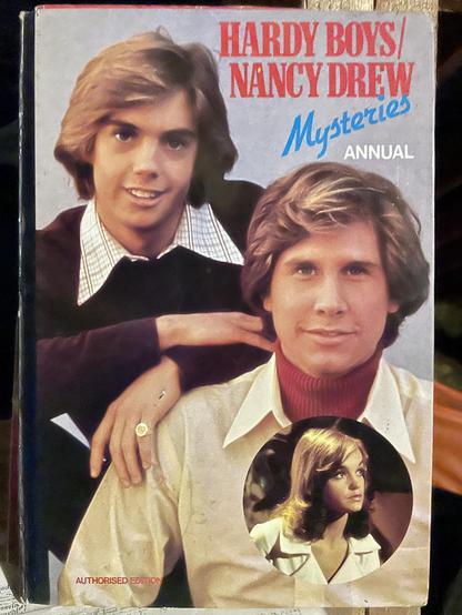 A vintage book cover featuring the title "Hardy Boys/Nancy Drew Mysteries Annual." Two young men are depicted smiling at the camera, with a smaller circular image of a young woman below. The men have stylish eighties hairstyles and are dressed in wide collared shirts