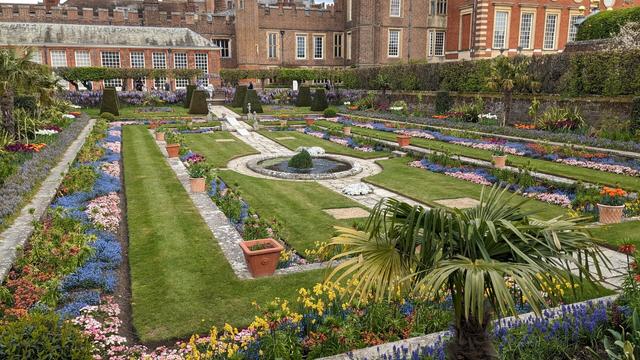 A formal garden with flower beds filled with plants of various bright colours. The grass is cut short and there's a stone path running through the garden, with a pond in the middle. In the background stands a brick building of several different eras, with both Tudor and Stuart sections of Hampton Court Palace visible.