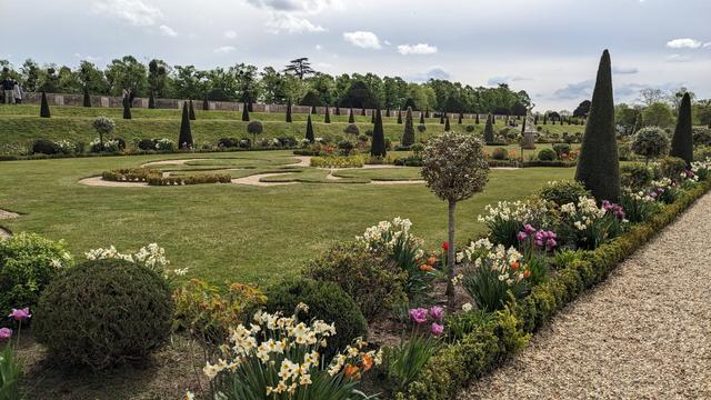 A vast formal garden with flower beds filled with plants of various bright colours. The grass is cut short and there are several well-kept topiaried bushes.