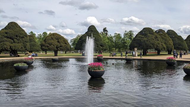 In the foreground is a large human-made pond, in the centre of which is a fountain with several jets. Surrounding this and evenly-spaced in the water are eight planters filled with flowers, alternating between pink and greenish. In the background are lawns bordered by large trees. Many people are walking around or sitting on benches.