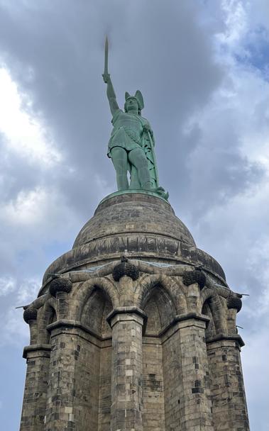 A tall statue of a historical figure in armor, holding a sword and raising it above their head. The statue is mounted on a circular stone pedestal with arched structures, set against a cloudy sky.