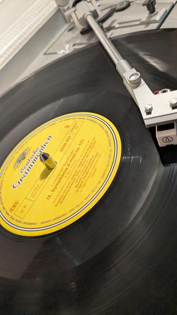 Das Bild zeigt eine Nahaufnahme einer laufenden Schallplatte auf einem Plattenspieler.

Auf dem gelben Label in der Mitte der Platte ist deutlich das Logo der Deutschen Grammophon zu erkennen.

Die Platte enthält Ludwig van Beethovens 9. Symphonie (d-Moll, op. 125), auch bekannt als die „Ode an die Freude“.

Der silberfarbene Tonarm des Plattenspielers ist abgesenkt, sodass die Nadel auf der Platte aufliegt und Musik abgespielt wird.

Die Platte dreht sich, was durch die leicht verschwommene Bewegung auf dem Foto sichtbar ist.

Die Oberfläche des Plattenspielers ist silberfarben, und das bekannte Audio-Technica-Logo ist in der Ecke zu sehen.

Dieses Bild fängt den Moment klassischen Musikgenusses im analogen Format ein – Beethoven aus der Rille, sozusagen.