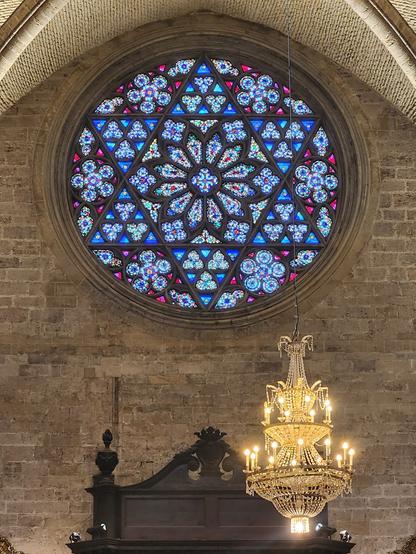 large round stained glass window with six-pointed star.  color scheme is mostly blue with background in mostly reds/purples. a chandelier hangs to its bottom right.
