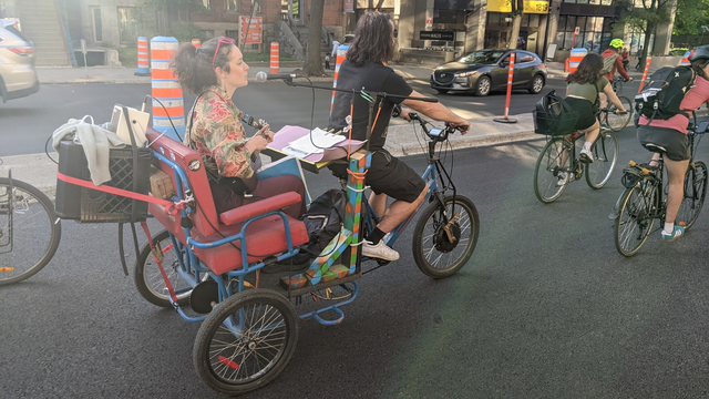 Photo du Cyclo-Pouce à l'été 2024, un vélo électrique à trois roues avec une banquette à l'arrière, roulent au milieu de la masse critique. Une guitariste chante et joue de la guitare à l'arrière. Un micro et ampli de guitare sont connectés à un haut-parleur attaché au dossier de la banquette, face aux cyclistes qui participent à la masse critique.