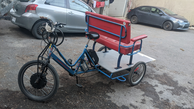 Photo du même Cyclo-Pouce dans une cours arrière après la modification - la banquette fait maintenant face à l'arrière afin de pouvoir voir les cyclistes, et la plateforme a été rallongée pour donner de la place au lutrin, ampli, haut-parleur et accessoires.