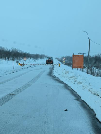 Snowy road with a barrier and a truck behind. An orange roadsign on the right is listing the convoy timetable.