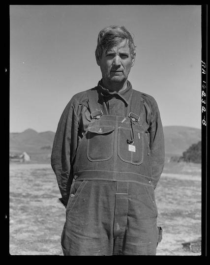 The image is a black and white photograph of an older man standing in what appears to be an open, flat field with mountains visible in the background. The sky is clear, indicating it might be taken during daytime. He has short gray hair, bushy eyebrows, deep-set eyes that gaze directly into the camera lens, and his facial expression looks pensive or serious.

He's wearing a pair of dark denim overalls which are baggy around the torso but fitted at the waist with visible pockets on both sides. The straps hanging from what seems to be suspenders are also made of cloth, not metal as is more commonly seen today for this purpose. A tag hangs off one strap near his right shoulder.

In the background, there's a small structure that could possibly be an outbuilding or part of farm equipment like a tractor parked at some distance away on the field. The overall setting suggests rural farmland work conditions typical in agricultural regions during what looks to be mid-20th-century America due to clothing style and lack of color imagery.

The photograph appears historical, likely taken as part of Dorothea Lange's documentation projects capturing subjects like migrant workers or tenant farmers which were prevalent themes in her works. The reference suggests it was captured from Texas who is now working in California during pea picking season at Nipomo, a region known for its agriculture activity.

A white label with text appears on t [...]