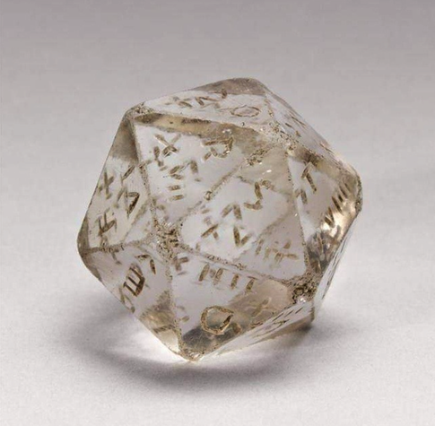 An clear icosahedron cut from crystal. There are three or more letters of the Roman alphabet on each face. The die is a bit worn from time and has some dirt caked in the carved letters.
