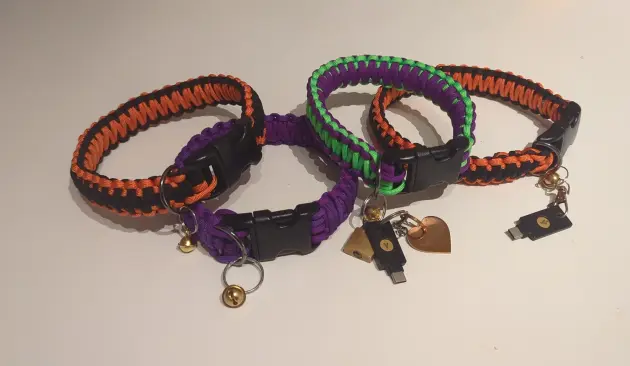 A photo of 4 paracord collars. Each of them are made out of different colors: orange, black, purple, neon green. They look really cool and have bells and Yubikeys attached to them.