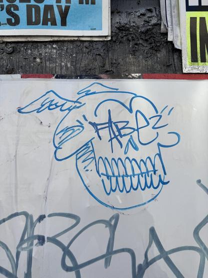 A drawing of a skull in a graffiti style on a wall in San Francisco. The skul has a subtle smile and wings coming out of the top of its head. Where the eyes would be, the artist has signed their name "Fabez". This is Fabez signature character, used throughout their tags and always with "Fabez" written over the eyes.