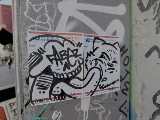 Two USPS mailing labels stuck onto a wall. The stickers have hand drawn art on them in a graffiti illustration style that shows a skull licking a cat. The skull looks happy, and the cat looks grumpy. The skull is mostly on one sticker and has a signature by "Fabez" on it. The cat is on another sticker and has a signature by "Phobic".