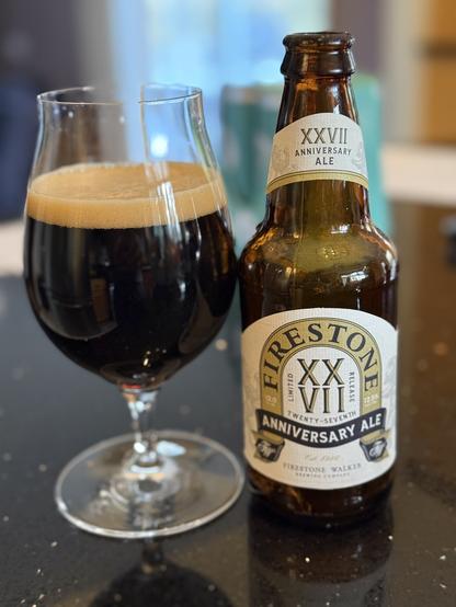 A snifter full of dark beer with a thin tan head. The brown bottle has white body and neck labels with “XXVII” on the label center