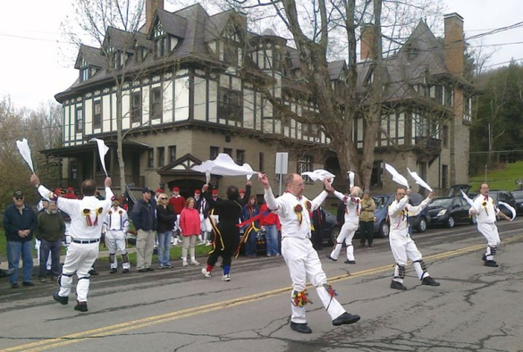 Six men (five dressed in white clothing and one in black, decorated with colored ribbons, with bells tied to their shins and white handkerchiefs in their hands) dance in a street in front of a large tudor revival inn. A crowd watches from the curb.