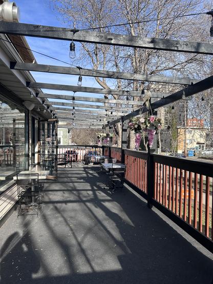 An empty but spacious patio with a few small tables for 2 people and a bench at the far end. Outdoor string lights are suspended from widely spaced black-painted wood beams above the seating area, and a couple of pink flower arrangements also hang from these. A wooden railing separates the raised patio from the lawn and sidewalk below.