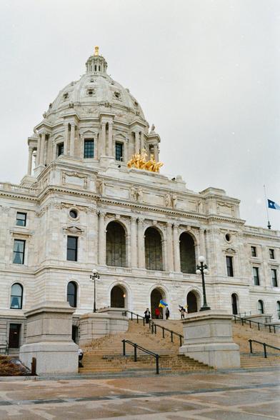 A view of the Minnesota State Capitol building on a cloudy day, with its large white dome and golden chariot sculpture prominently visible. On the front steps, a small group of people hold Ukrainian flags, showing support for Ukraine.