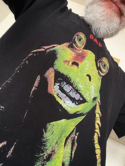 A person is wearing a black T-shirt featuring a colorful graphic of Jar Jar Binks a fictional alien from the Star Wars universe. The person has a white beard, and part of their arm is visible in the frame.