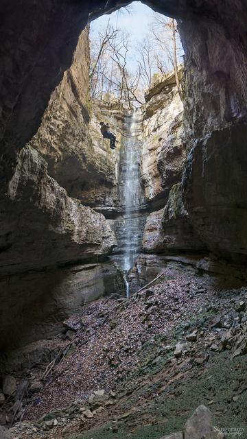 A vertical pit cave as a vertical panoramic. The top is blue skies above with trees stretching up towards the heavens. A waterfall in the back beats its way into the depths of the pit, full of moss and leaves. A climber is on rope just left of the waterfall, hanging in freespace, but making their way towards the light.