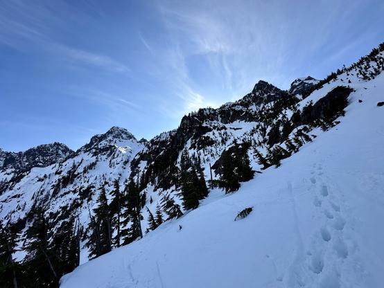 A winter landscape featuring snow-covered mountains under a clear blue sky, with pine trees in the foreground and a hint of sunlight emerging from behind the peaks. Snow track steps are visible on the ground.