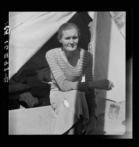 The image is a black and white photograph of an elderly woman seated on the edge of what appears to be a wooden platform. She wears a knitted sweater with stripes in shades of gray, beige, and cream. The texture suggests it may have been worn for some time or possibly patched due to wear.

She has pale skin, visible wrinkles around her eyes and mouth indicating age, and is looking directly at the camera with an expression that could be interpreted as contemplative or weary. Her hair is thinning on top of her head but remains combed back neatly from it appears she is balding in some areas.

The woman's right arm rests casually on a wooden surface while her left hand holds what seems to be a small object, perhaps a piece of bread. A tin can sits near the edge beside her, empty and tilted slightly towards herself as if she has just set it down.

Behind her is an open tent or shelter with fabric walls that appear tattered in places due to wear or possibly wind exposure. The interior reveals another individual partially visible under the cover, resting on a makeshift bed composed of what looks like burlap sacks filled with straw or similar material. This setting suggests a temporary living situation.

The shadow cast by her and part of the tent indicates bright sunlight illuminating this scene from one side, casting defined shadows to the opposite direction.

Additional text overlaid at the top left corner reads "5H 1934" whi [...]