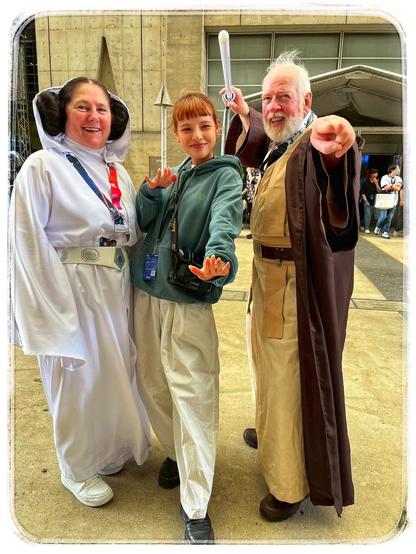 Three individuals pose together in costume, referencing a sci-fi theme. One person wears a Princess Leia costume, another is in casual clothing with a hoodie, and the third is dressed as a Jedi, holding a lightsaber. They are smiling and standing outside