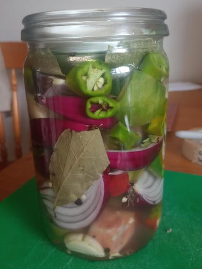 A jar full of roughly chopped bright green and red chilles, purple onion and green tomato with a reddening centre. The jar has bayleaves, pepper and a vine leaf on top.