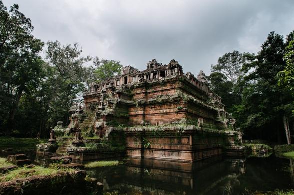 Phimeanakas, a Hindu Temple in Siem Reap, Cambodia stands among trees and surrounded by water. The temple is an orangish red colour.