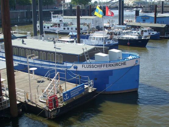 Ein altes Binnenschiff mit blauem Rumpf im Hafen. Brauner Aufbau mit Rundbogenfenstern. Am Bug auf weißem Grund der Name: "Flussschifferkirche".