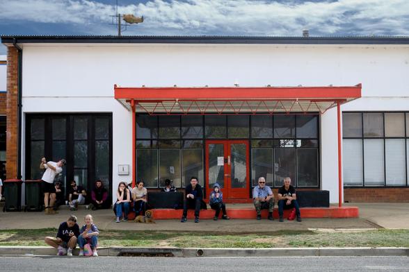 Photo of spectators sitting in front of a building with a prominent red awning, above which floats a plastic fish.