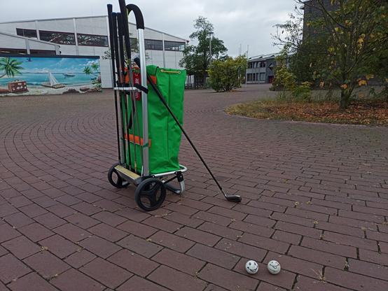 Auf einem Svhulhofsgelände steht ein grasgrüner Hackenporsche, an dem Golfschläger befestigt ein.
Davon liegen zwei weiße Golfbälle mit Smiliegesicht.
Der Boden ist mit roten Steinen gepflastert