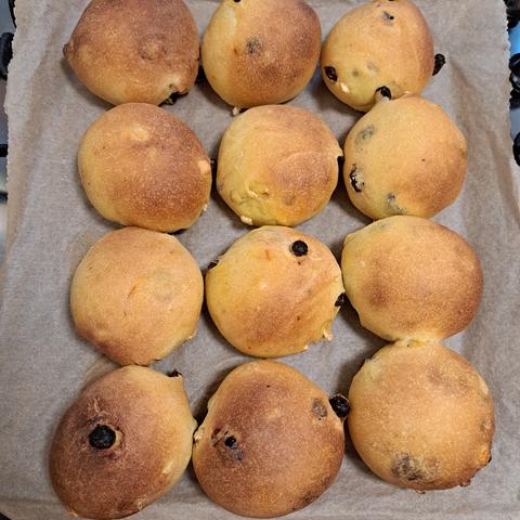 Twelve golden coloured buns on a baking sheet lined with paper. The raisins and candid peel can be seen poking out of some of the buns. With others their sweetness  an be seen just below the skin.