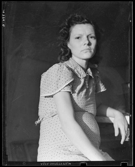 The image is a black and white photograph featuring a pregnant woman sitting with her body slightly turned to the side. She appears contemplative or possibly fatigued, as she gazes off into space with a neutral expression on her face.
She wears a short-sleeved dress adorned with small dots and has puffed sleeves at the shoulders. The pattern of the dress suggests it may be from an earlier era, potentially mid-20th century based on its style. Her hair is styled in loose waves that fall around her shoulder area. No discernible background elements can be seen; she appears to be sitting against a plain backdrop or possibly seated at a table with nothing immediately notable within the frame of the photo.
This image carries historical context, as indicated by the text and metadata provided which suggests it's part of Dorothea Lange’s documentation on migrant families in Imperial Valley, California. The photograph captures a moment from the life of this woman who is presumably pregnant at the time due to her visible baby bump.