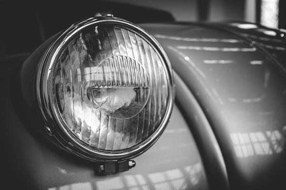 Close-up black and white photo of a vintage car headlight on display in a museum. The curved bodywork and polished chrome frame reflect the overhead lights and surroundings, emphasizing the classic design and craftsmanship.