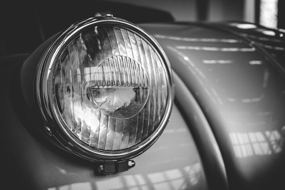 Close-up black and white photo of a vintage car headlight on display in a museum. The curved bodywork and polished chrome frame reflect the overhead lights and surroundings, emphasizing the classic design and craftsmanship.