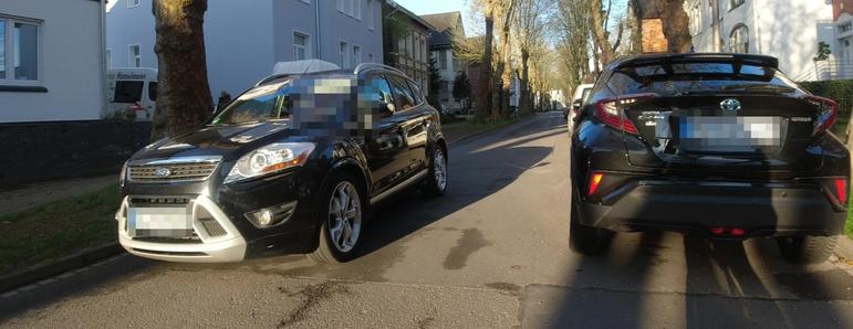 Sicht aus einer leicht außermittig am Lenker eines Fahrrades montierten Kamera. In Fahrtrichtung rechts parkt ein KFZ, weswegen der Fahrradfahrer wartet, bis ein entgegenkommendes KFZ passiert hat. Der verbleibende Platz zwischen parkenden KFZ und Gegenverkehr ist zu schmal für einen Menschen auf dem Fahrrad.
