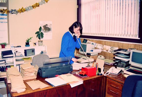 A 1980's office, dark wooden desks piled with envelopes and other paperwork. A lady in a blue knitted dress answers the phone taking notes with pen and paper, she has both a Brother electronic typewriter and a BBC Micro computer.
