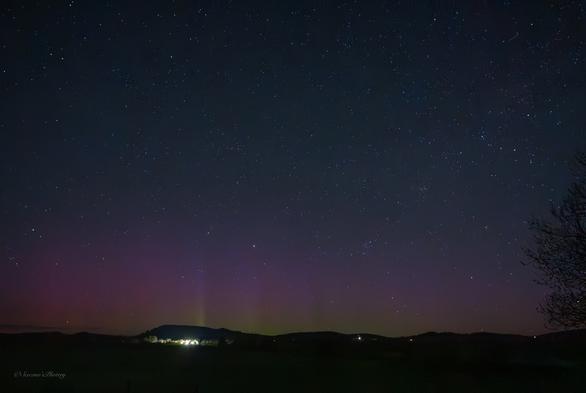 A dark, early morning sky filled with stars above a hilly horizon. Directly above the horizon are streaks of bright green into red Aurora. The lights of houses are scattered along the horizon