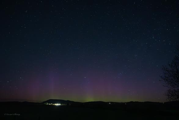 A dark, early morning sky filled with stars above a hilly horizon. Directly above the horizon are streaks of bright green into red Aurora. The lights of houses are scattered along the horizon