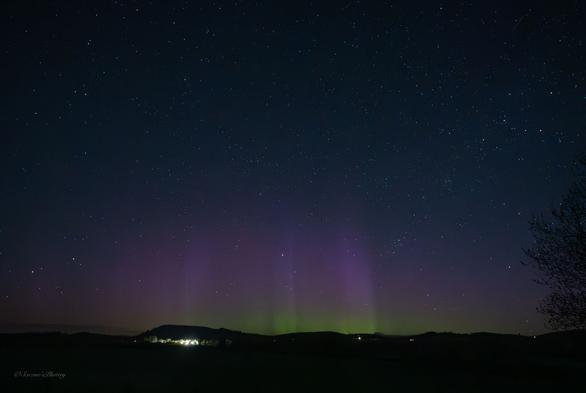 A dark, early morning sky filled with stars above a hilly horizon. Directly above the horizon are streaks of bright green into red Aurora. The lights of houses are scattered along the horizon