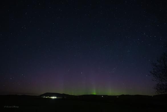 A dark, early morning sky filled with stars above a hilly horizon. Directly above the horizon are streaks of bright green into red Aurora. The lights of houses are scattered along the horizon