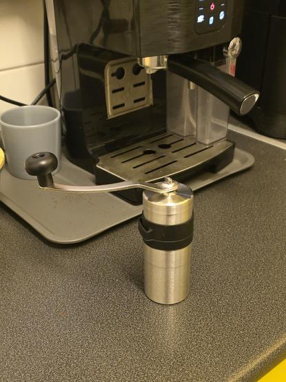 The image shows a kitchen countertop with a coffee machine and a manual coffee grinder. The coffee machine is black with a digital display and a portafilter handle, positioned on a gray tray. To the left of the machine, there is a gray mug. In the foreground, a stainless steel manual coffee grinder with a black grip and handle is prominently displayed. The countertop is dark gray with a speckled texture. The background includes a white tiled wall and a yellow lower cabinet edge.

Provided by @altbot, generated privately and locally using Ovis2-8B

🌱 Energy used: 0.137 Wh