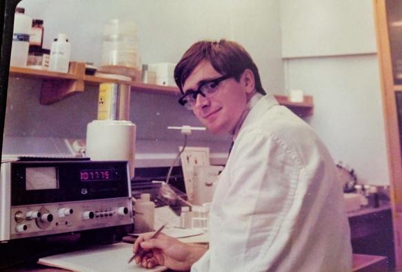 Man wearing a white coat holding a pen is in front of a device with numbers showing. There is a white container with a metal box above it with a radioactivity symbol. There are shelves on the wall. The man has strange hair and even stranger glasses. He's trying hard not to laugh at the way he looks.