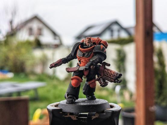 Pomalowana figurka Space Marine z Warhammer 40,000 w dynamicznej pozie, w barwach pomarańczowo-czarnych.  Marine trzyma w prawej ręce pistolet, a w lewej miecz częściowo dobyty z pochwy. Przy biodrze wisi miotacz płomieni. Zbroja jest głównie czarna z wyraźnymi pomarańczowymi elementami na plecaku, napierśniku, nakolannikach i naramiennikach. Figurka stoi na okrągłej podstawce, sfotografowana na tle rozmytego ogrodu i domów.