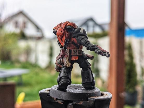 Tył pomalowanej figurki Space Marine w barwach pomarańczowo-czarnych. Na zdjęciu widoczny jest szczegółowo plecak z pomarańczowymi akcentami, przytroczone sakwy oraz detale ekwipunku. Marine trzyma broń w prawej ręce, a całość ustawiona jest na okrągłej podstawce, z rozmytym ogrodem i domami w tle.